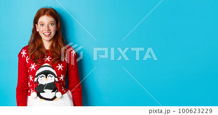 Winter holidays and Christmas Eve concept. Excited redhead girl in xmas sweater looking surprised at camera, standing against blue background 100623229