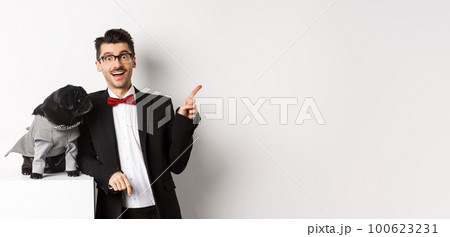 Animals, party and celebration concept. Happy and amazed young man and cute pug in costume standing over white background, dog owner pointing right Animals, party and celebration concept. Happy and amazed young man and cute pug in costume standing over white background, dog owner pointing right 100623231