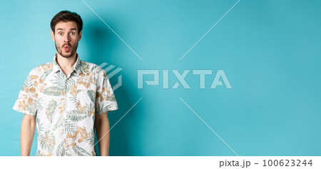 Summer holiday. Surprised tourist say wow and staring at camera, checking out awesome promo, standing in hawaiian shirt on blue background 100623244