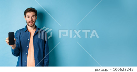 Happy caucasian guy showing empty mobile screen and smiling, standing on blue background 100623245