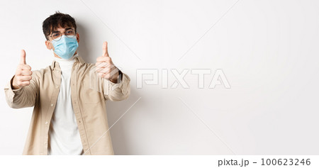 Coronavirus, health and real people concept. Smiling guy in medical mask showing thumbs up, wearing glasses, standing on white background 100623246