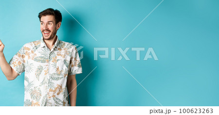 Excited smiling tourist pointing and looking aside at empty space, checking out promo offer, standing in hawaiian shirt on blue background 100623263