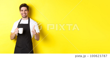 Barista bringing coffee and pointing finger gun at camera, standing in black apron against yellow background Barista bringing coffee and pointing finger gun at camera, standing in black apron against yellow background 100623787