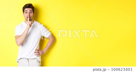 Image of excited guy planning surprise, shushing at camera, press finger to lip in hush sign, standing over yellow background 100623801