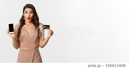 Online shopping and holidays concept. Excited beautiful lady in elegant dress showing mobile screen and credit card, standing over white background 100623806