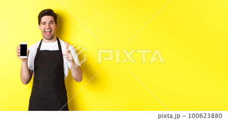 Happy waiter showing mobile screen and thumb up, recommending cafe restaurant app, standing over yellow background Happy waiter showing mobile screen and thumb up, recommending cafe restaurant app, standing over yellow background 100623880