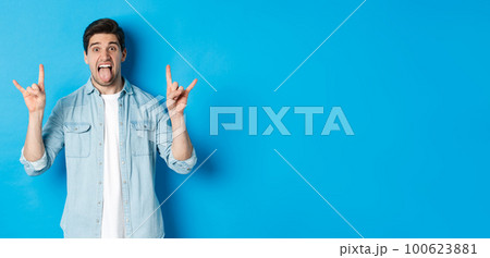 Cool guy having fun, enjoying concert, showing rock sign and tongue, standing over blue background Cool guy having fun, enjoying concert, showing rock sign and tongue, standing over blue background 100623881
