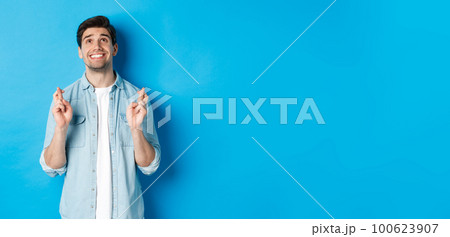Lucky guy praying and making wish with crossed fingers, looking up with pleading face, standing against blue background 100623907