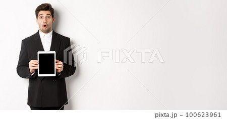 Image of surprised handsome man in black suit, showing digital tablet screen and looking amazed, standing against white background 100623961