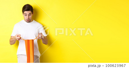 Surprised boyfriend open bag with gift, looking inside amazed, standing over yellow background Surprised boyfriend open bag with gift, looking inside amazed, standing over yellow background 100623967