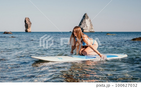 Woman sea sup. Close up portrait of happy young caucasian woman with long hair looking at camera and smiling. Cute woman portrait in a blue bikini posing on sup board in the sea 100627032