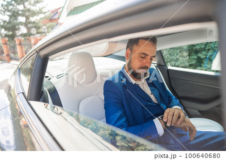 A caucasian man in a blue suit looks at his watch while sitting in the back seat of a car. Business class passenger. A caucasian man in a blue suit looks at his watch while sitting in the back seat of a car. Business class passenger. 100640680