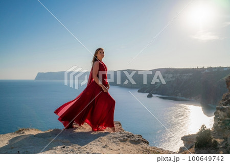 A woman in a red flying dress fluttering in the wind, against the backdrop of the sea. A woman in a red flying dress fluttering in the wind, against the backdrop of the sea. 100649742