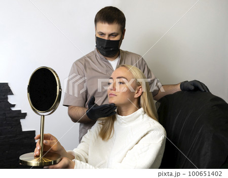 Doctor, beauty physician shows woman results of beauty injection procedure, face reshape with filler. White smiling happy female looks at mirror. Lifestyle, Aesthetic medicine. Horizontal plane 100651402