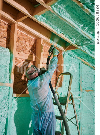 Male builder insulating wooden frame house. Man worker spraying polyurethane foam inside of future cottage, standing on ladder, using plural component gun. Construction and insulation concept. Male builder insulating wooden frame house. Man worker spraying polyurethane foam inside of future cottage, standing on ladder, using plural component gun. Construction and insulation concept. 100659234