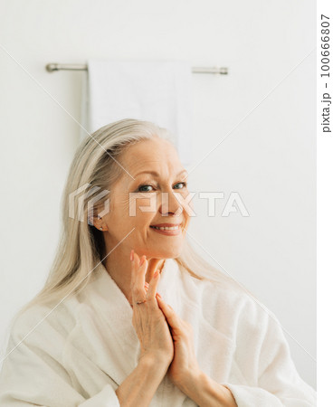 Smiling female with grey hair admires her reflection while standing in front of a bathroom mirror in the morning	 100666807