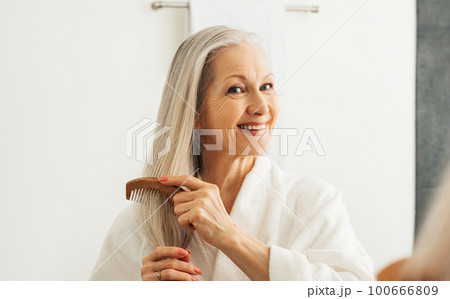 Cheerful aged female combing her gray hair in front of a bathroom mirror	 100666809