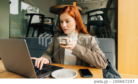 Woman working as a digital nomad in a cafe. Female entrepreneur with ginger hair holding a cup typing on a laptop Woman working as a digital nomad in a cafe. Female entrepreneur with ginger hair holding a cup typing on a laptop 100667297