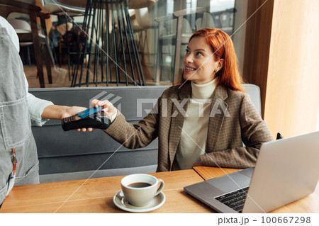 Smiling businesswoman with ginger hair paying by card in coffee shop Smiling businesswoman with ginger hair paying by card in coffee shop 100667298