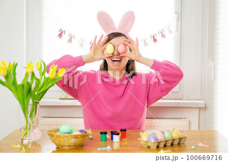 Happy beautiful young woman smiling wearing rabbit ears holding Easter eggs for her eyes. 100669716