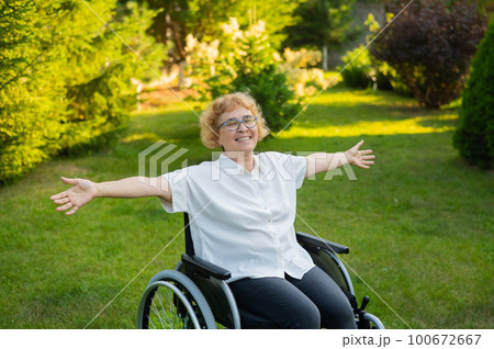 Happy old woman spread her arms to the sides while sitting in a wheelchair on a walk outdoors.  100672667