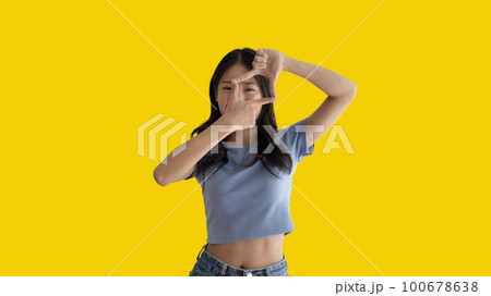Playful young asian woman in good mood posing for photo or photo frame with fingers on yellow background,  Use your fingers to act like a photograph, Mark as if taking a photo. 100678638
