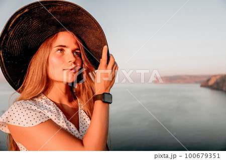 Portrait of happy young woman wearing summer black hat with large brim at beach on sunset. Closeup face of attractive girl with black straw hat. Happy young woman smiling and looking at camera at sea. Portrait of happy young woman wearing summer black hat with large brim at beach on sunset. Closeup face of attractive girl with black straw hat. Happy young woman smiling and looking at camera at sea. 100679351