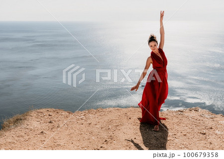 Woman in red dress on sea. Side view a Young beautiful sensual woman in a red long dress posing on a rock high above the sea on sunset. Girl on the nature on blue sky background. Fashion photo. Woman in red dress on sea. Side view a Young beautiful sensual woman in a red long dress posing on a rock high above the sea on sunset. Girl on the nature on blue sky background. Fashion photo. 100679358