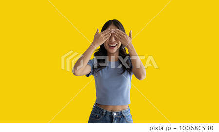 Young woman covering her face and eyes with her hands isolated, Cover your eyes with your hands for fear or prying eyes, Sneak peek, Yellow background. 100680530