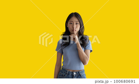 Asian woman doing silent gesture with finger, Noiseless symbol, Noisy mall, Hide a secret, Do not speak. shut your mouth, Noiseless signal transmission, Isolated on yellow background. 100680691