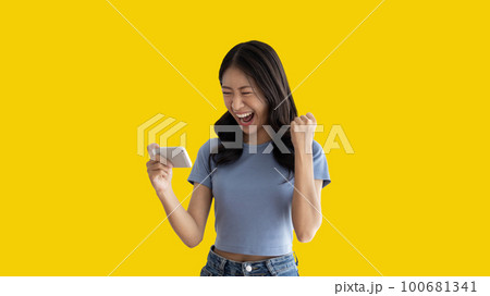 Asian woman playing games on mobile phone and wearing headphones having fun, Play games on the sofa in the living room on weekends, Resting at home, Comfort zone, Touch screen mobile phone. 100681341