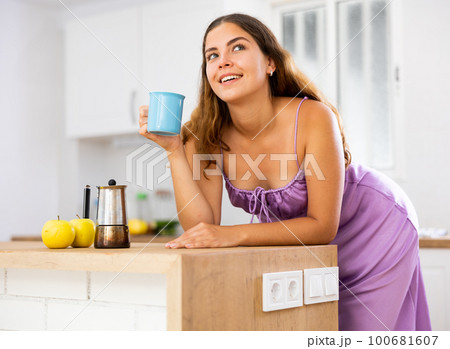 Portrait of young woman leaning on table in kitchen and drinking coffee Portrait of young woman leaning on table in kitchen and drinking coffee 100681607