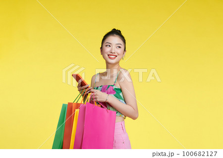 Young excited cheerful woman holding shopping bags isolated over yellow background using mobile phone looking aside. 100682127