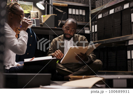 Law enforcement agents sitting on flooor and investigating crime together. African american man and woman detective partners analyzing case files and drinking coffee at night time 100689931