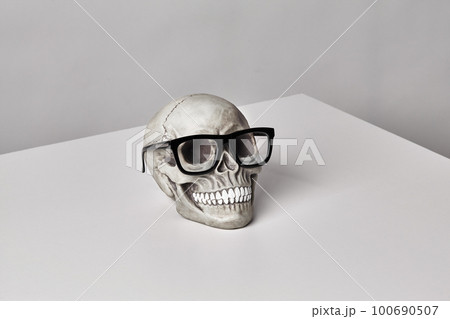 Realistic model of a human skull with teeth in a black-framed classic glasses on a light table, white background. Halloween horror concept. 100690507
