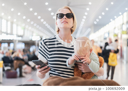 Mother traveling with child, holding his infant baby boy at airport terminal, checking flight schedule, waiting to board a plane. Travel with kids concept 100692009