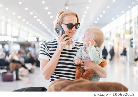 Mother talking on mobile phone while traveling with child, holding his infant baby boy at airport terminal waiting to board a plane. Travel with kids concept 100692018