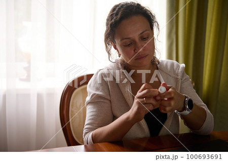 Overworked multi-ethnic business woman, freelancer entrepreneur sitting at desk with laptop and holding eye drops in her hands. Person with dry vision overworking suffering from digital eye strain 100693691