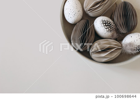 Moody spring still life scene. Easter composition. Bowl of folded paper ornaments, hen eggs decorated by guineafowl bird feather. Beige table background. Neutral festive flat lay, top view. Web banner 100698644