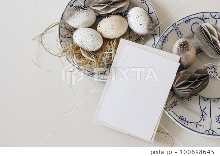 Easter stationery still life. Blank greeting card mockup. Blue plates with traditional speckled eggs decoration with feather. Beige paper ornaments. Spring flat lay, top view, holiday bannner. 100698645