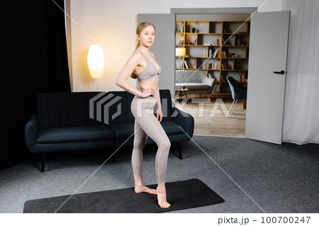 A cute slender girl is preparing for yoga training. Girl standing on yoga mat at home in living room A cute slender girl is preparing for yoga training. Girl standing on yoga mat at home in living room 100700247