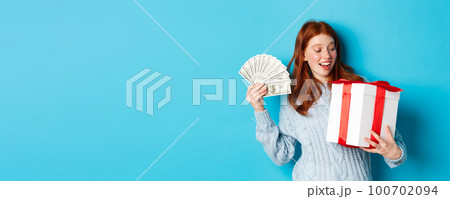 Christmas and shopping concept. Cheerful girl with red hair, holding money and big New Year gift, smiling happy, standing over blue background 100702094