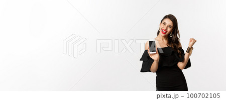 Online shopping concept. Satisfied pretty woman showing smartphone screen, making fist pump to rejoice, winning in internet, standing over white background 100702105