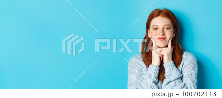 Close-up of cute and silly girl with red hair poking cheeks, showing dimples and smiling, standing against blue background 100702113