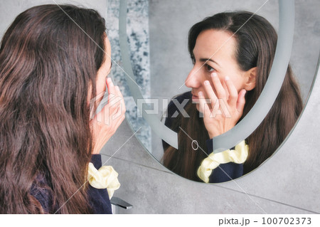 An attractive white Caucasian young woman looking in the bathroom mirror touching her wrinkles around her eyes An attractive white Caucasian young woman looking in the bathroom mirror touching her wrinkles around her eyes 100702373