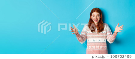 Image of happy redhead girl holding big size object, looking amazed and smiling, receiving Christmas gift, standing over blue background 100702409
