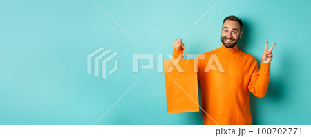 Happy young man showing peace sign and orange shopping bag, smiling pleased, standing over turquoise background Happy young man showing peace sign and orange shopping bag, smiling pleased, standing over turquoise background 100702771