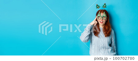 Christmas party and celebration concept. Cute redhead teen girl celebrating New Year, wearing xmas tree headband and funny glasses, looking left amused, blue background 100702780