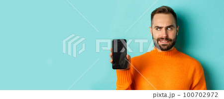 Confused guy scartching head and showing smartphone screen, looking puzzled, standing over turquoise background 100702972