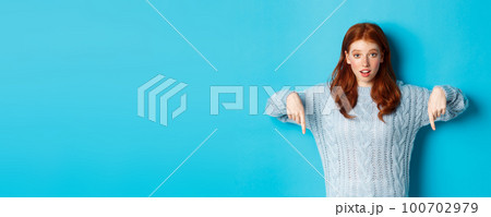 Winter holidays and people concept. Cute teenage girl pointing fingers down, showing advertisement, standing over blue background 100702979